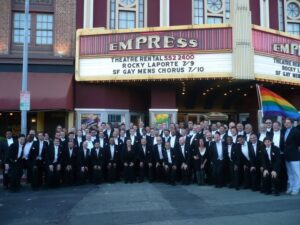 SFGMC singers in black tuxedos pose outside the Empress Theatre in Vallejo, its marquee announcing the San Francisco Gay Men’s Chorus.